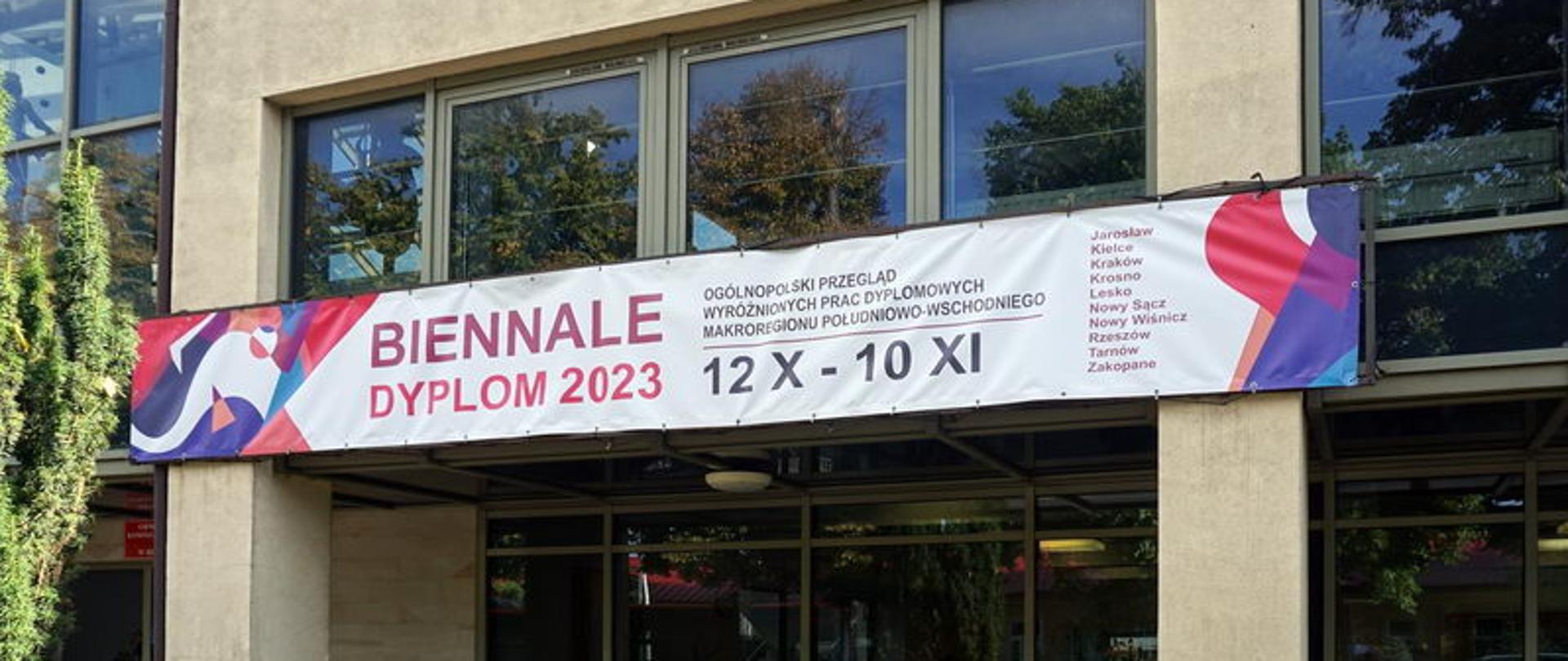 zdjęcie przedstawia wejście do szkoły nad którym zawieszony jest banner z napisem w kolorze czerwonym o treści "biennale dyplom 2023"