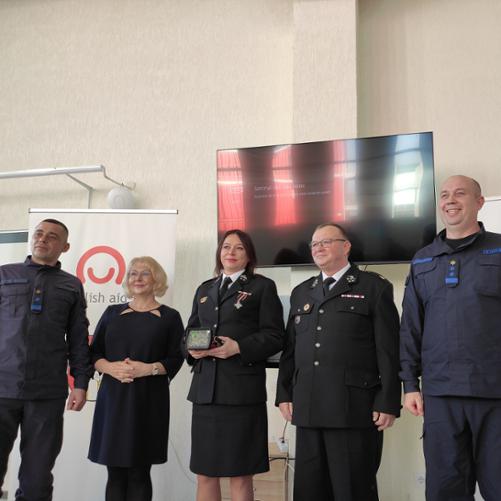 Do zdjęcia pozują dwie kobiety oraz trzech mężczyzn stojących przed ekranem i banerami z logo Polskiej pomocy; kobieta stojąca w środku grupy trzyma w rękach kamerę termowizyjną