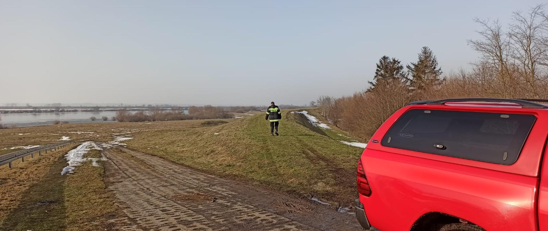 na wale przeciwpowodziowym stoi funkcjonariusz. W tle widać rzekę Wisłę po lewej tronie , po prawej drzewa i krzaki.