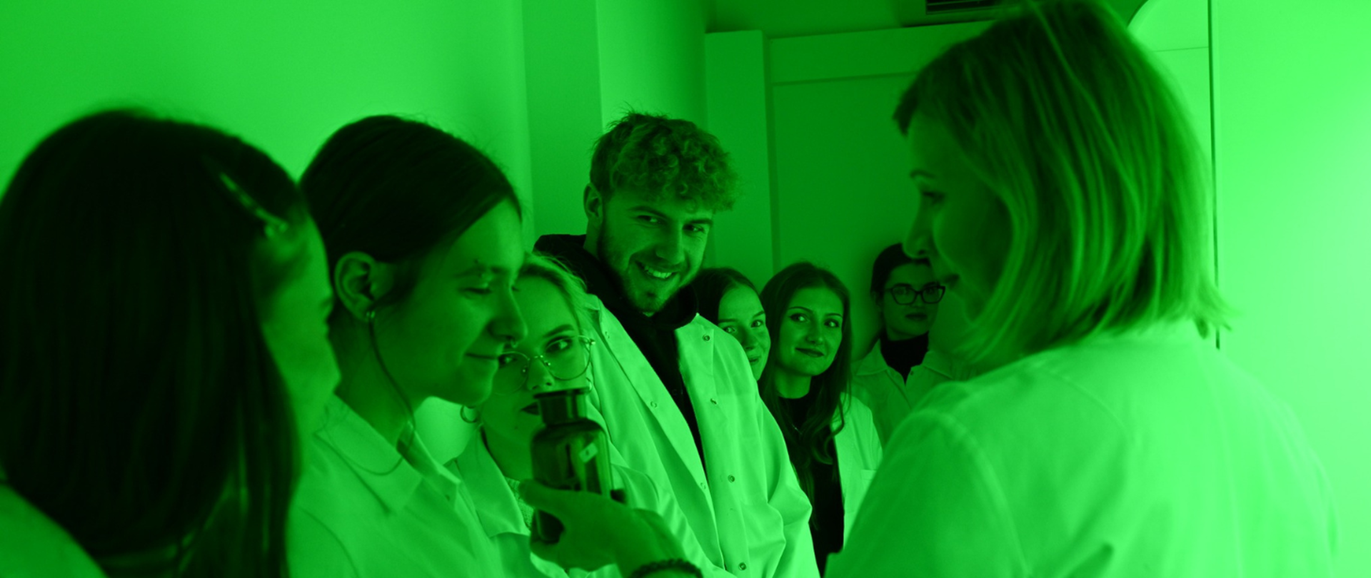 Studenci w białych fartuchach laboratoryjnych słuchają prowadzącej, która pokazuje im próbkę w laboratorium o zielonym oświetleniu.
