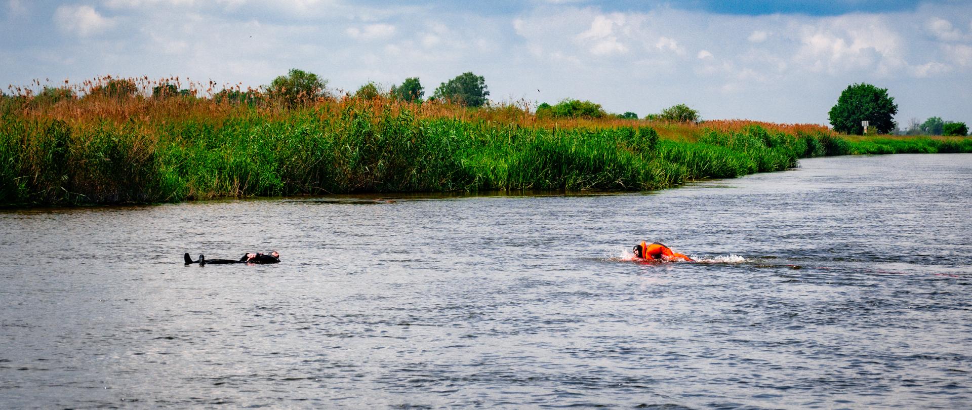 na rzece jedna osoba płynie w ubraniu pomarańczowym druga płynie bezwładnie w czarnym ubraniu w oddali zarośla