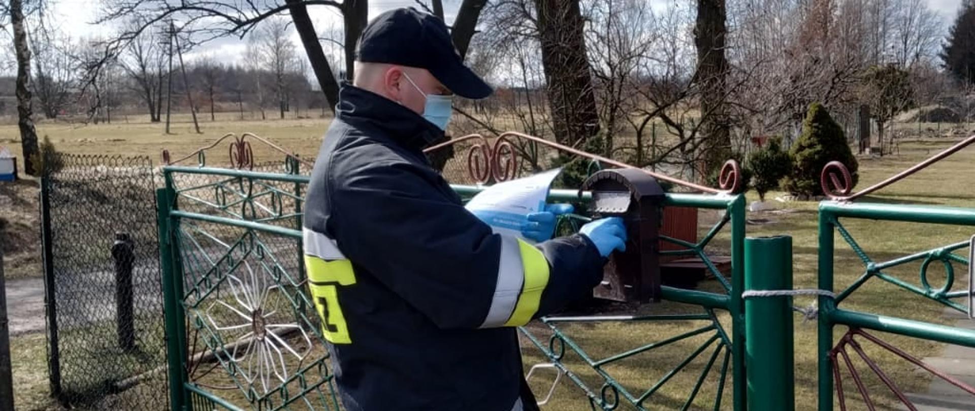 Zdjęcie przedstawia strażaka ochotniczej straży pożarnej ubranego w ubranie specjalne, wkładającego ulotkę do skrzynki pocztowej umieszczonej na ogrodzeniu posesji. Strażak ubrany w maseczkę i rękawiczki jednorazowe. W tle teren posesji. 