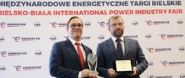 Minister Grzegorz Puda i przedstawiciel firmy ZPUE S.A. nagrodzonej nagrodą ministra funduszy i polityki regionalnej za innowacyjny produkt TPM Air – rozdzielnicę pierścieniowa średniego napięcia, minister trzyma w dłoniach tabliczkę informującą o nagrodzie, po lewej od niego przedstawiciel firmy ZPUE s.A. trzyma statuetkę z nagrodą