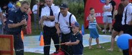 Na zdjęciu widać komentującego policjanta oraz strażaków z dzieckiem, który trzyma wąż od hydronetki. W tle festyn kręci mnie bezpieczeństwo. 