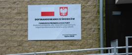 Tablica informacyjna na ceglanej ścianie budynku z polską flagą i godłem. Napis informuje o dofinansowaniu z Funduszu Promocji Kultury na budowę podjazdu dla osób z niepełnosprawnościami i zagospodarowanie terenu przy szkole; data podpisania umowy: lipiec 2025. Na pierwszym planie metalowy podjazd z poręczami.