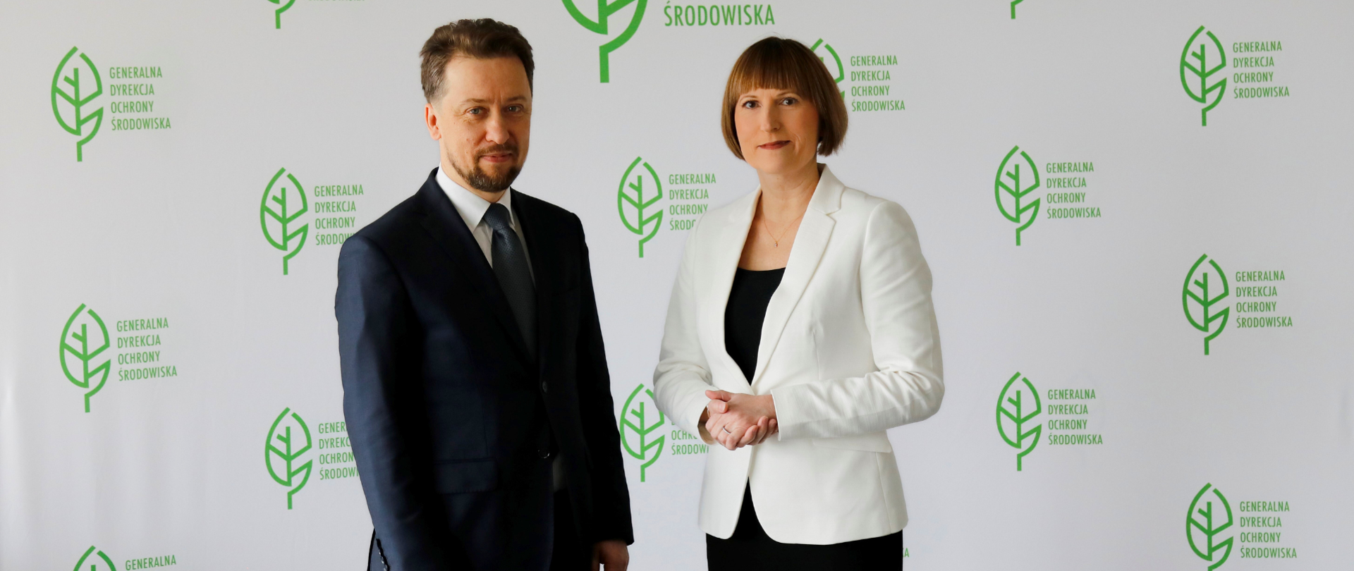 Na tle białej ściany z logotypem Generalnej Dyrekcji Ochrony Środowiska (zielony liść) stoi mężczyzna w garniturze i kobieta. Kobieta ubrana jest w czarną sukienkę oraz biały żakiet.