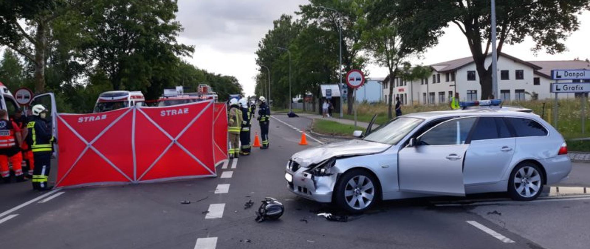 Na zdjęciu z lewej strony jest rozstawiony czerwony parawan z napisem "straż". Obok parawanu stoją strażacy oraz Zespół Ratownictwa Medycznego. Z prawej strony znajduje się srebrny samochód marki BMW. Samochód ma wgnieciony lewy przód. Przed samochodem leży kask motocyklisty. 