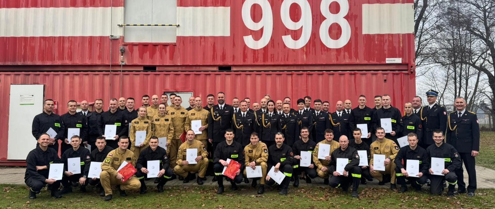 Pamiątkowe zdjęcie grupowe funkcjonariuszy PSP kończących szkolenie podstawowe w zawodzie strażak w towarzystwie Zastępcy Dolnośląskiego Komendanta Wojewódzkiego PSP, Zastępcy Komendanta Centralnej Szkoły PSP oraz innych funkcjonariuszy PSP przybyłych na uroczystość zakończenia szkolenia, w tle kontener do zajęć z pożarów wewnętrznych