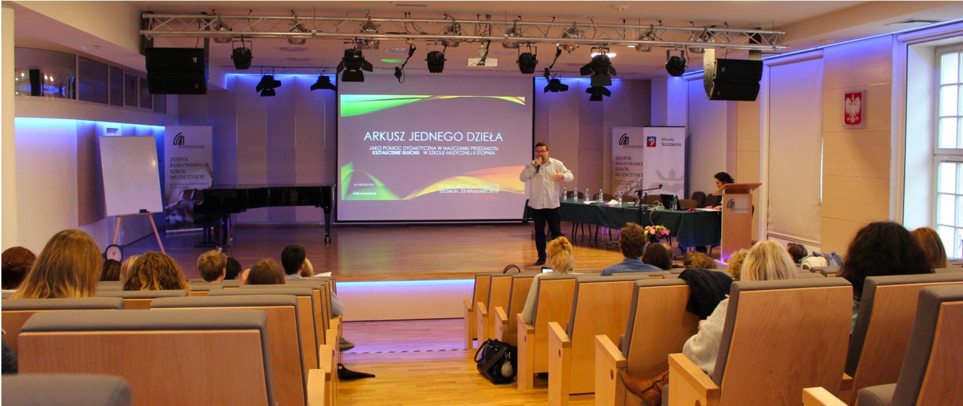 Grafika przedstawia wystąpienie Michała Moca prezentującego arkusz jednego dzieła na jednej z edycji konferencji