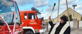 Na zdjęciu widać pojazd ratowniczo-gaśniczy na podwoziu Man z zabudową firmy Stolarczyk. Pojazd czerwony. Posiada białe oznaczenia. Posiada sygnalizację ostrzegawczą. Żaluzje zamknięte na zabudowie. W tle budynek OSP Rajsko. Pojazd stoi na asfalcie. Pora dnia to dzień. Przed pojazdem ksiądz dokonuje aktu poświęcenia.