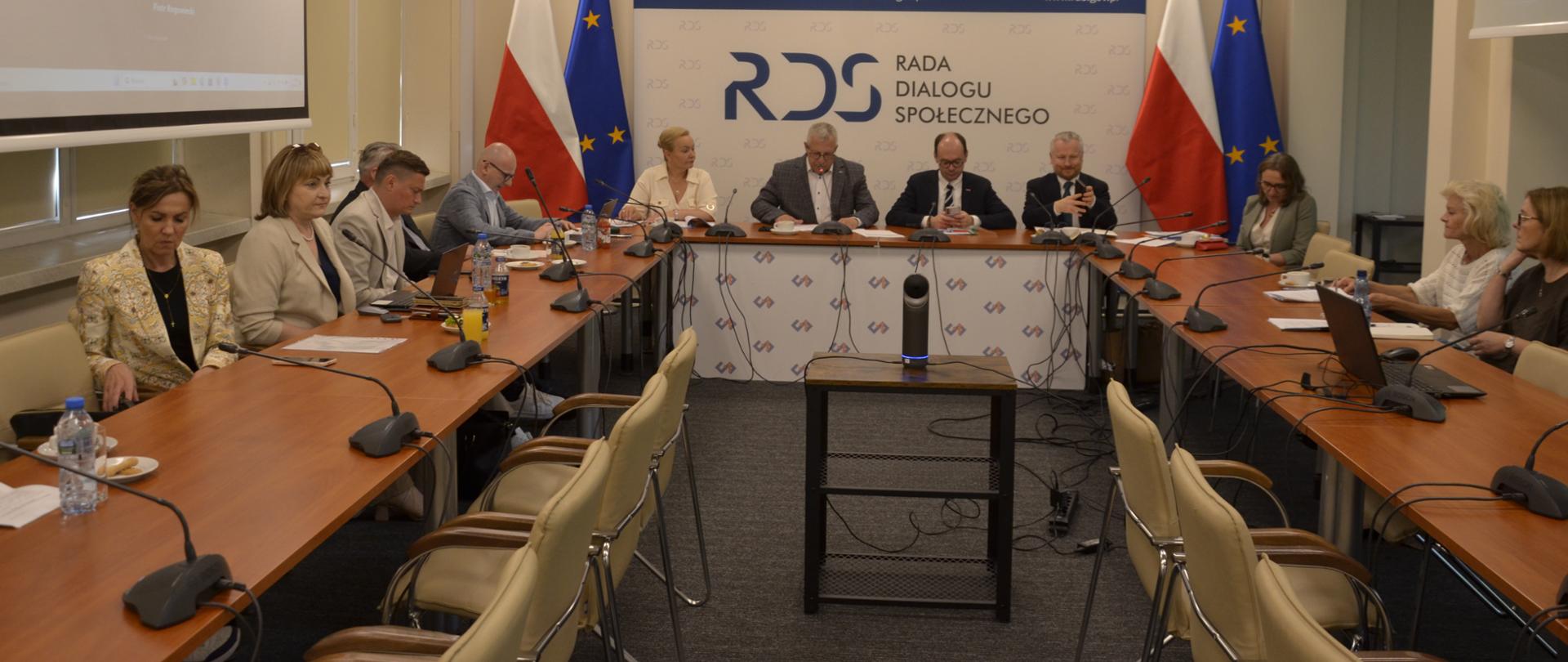 Na zdjęciu widnieje stół prezydialny za którym siedzą od lewej strony: Małgorzata Baranowska - Podsekretarz Stanu w Ministerstwie Rodziny, Pracy i Polityki Społecznej, Przewodniczący zespołu ds. budżetu, wynagrodzeń i świadczeń socjalnych – Leszek Miętek, Sebastian Gajewski - Podsekretarz Stanu w Ministerstwie Rodziny, Pracy i Polityki Społecznej Jurand Drop – Podsekretarz Stanu w Ministerstwie Finansów . Po obu stronach stołu widnieją dwie flagi: Polski i UE. W tle siedzących znajduje się baner z napisem Rada Dialogu Społecznego a nad nim wisi godło Polski. Ponadto pozostali uczestnicy spotkania