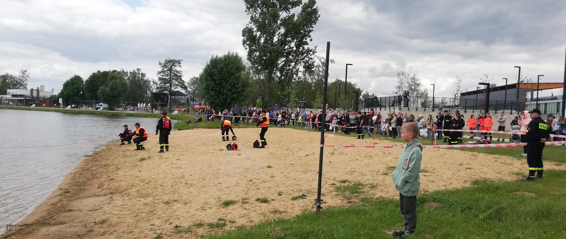 Na plaży przy jeziorze strażacy realizują pokazy ratownictwa wodnego. Wokół plaży zebrane grono obserwatorów uczestniczących w wielkim otwarciu. Strażacy ubrani w kamizelki ratunkowe prowadzą działania ratunkowe na linii brzegowej asekurując rotę pracującą w wodzie.