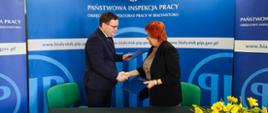 2 osoby na tle banneru w kolorze niebieskim z logo i napisem Państwowa Inspekcja Pracy. Osoby trzymają teczki w kolorze niebieskim i wymieniają uścisk dłoni w geście zawarcia umowy. Od lewej strony: Pan dr Karol Łapiński p.o. Okręgowego Inspektora Pracy w Białymstoku, Pani dr n. med. Maria Siewko – Podlaski Państwowy Wojewódzki Inspektor Sanitarny w Białymstoku.