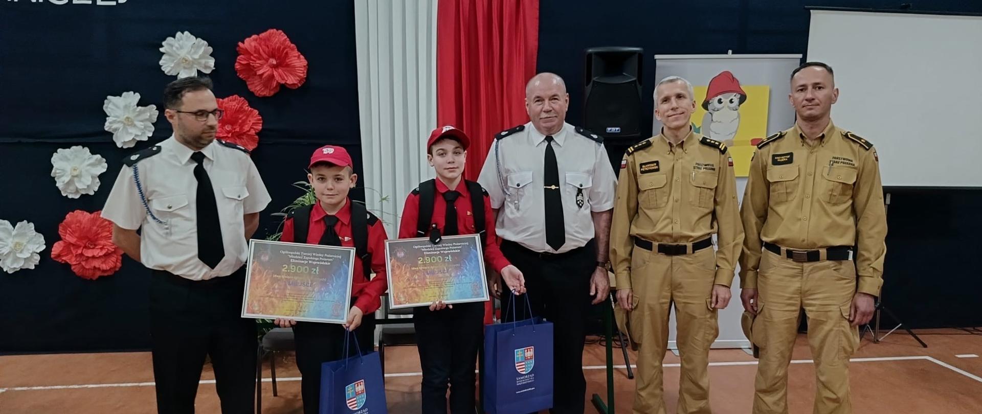 Grupa ludzi na tle flagi Polski. 4 dorosłe osoby wręczają nagrody dla 2 młodych strażaków. 