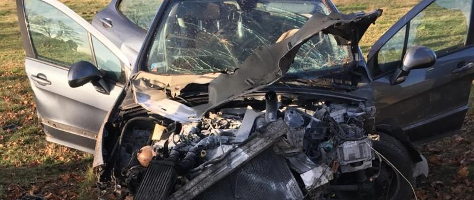 Na fotografii widać rozbity samochód w kolorze czarnym. Samochód ma rozbity przód oraz otwarte drzwi. Znajduje się na poboczu na trawie. Wokół pojazdu widać oderwane elementy konstrukcyjne. W tle widać łąki i granicę lasu.