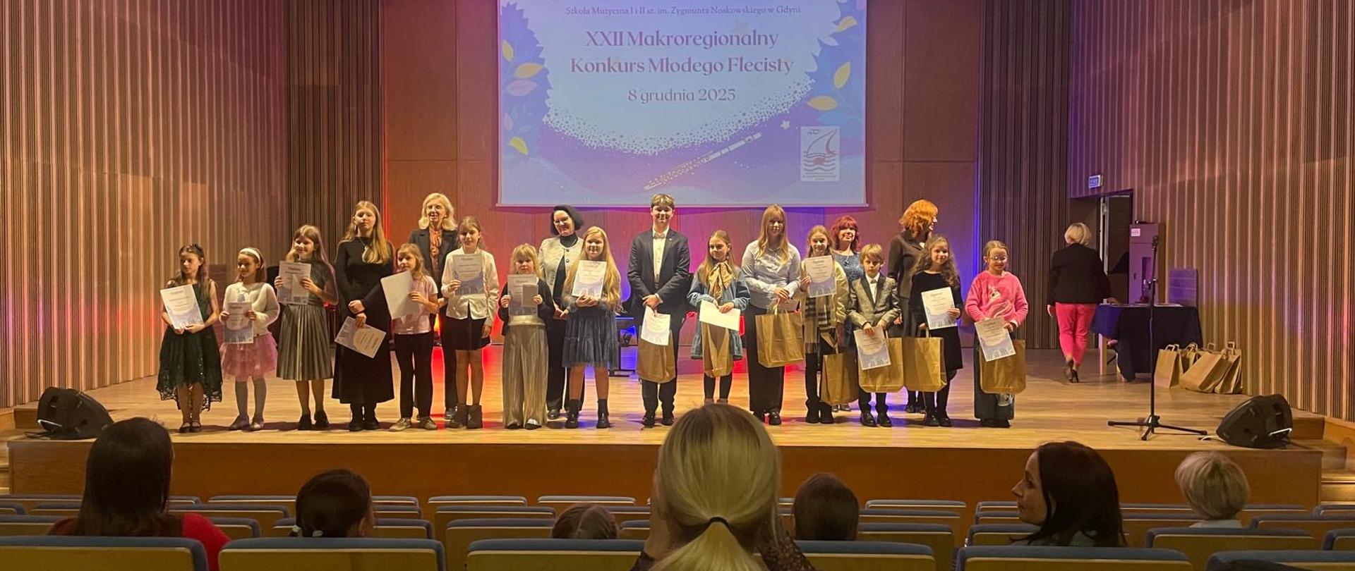 Na scenie stoją nagrodzeni uczestnicy konkursu razem ze swoimi opiekunami. W tle na ekranie wyświetlony jest plakat konkursu. 