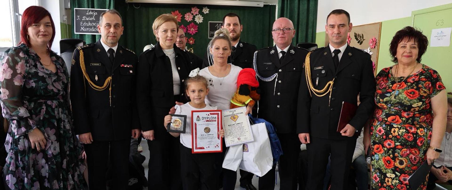 Na zdjęciu widać grupę ośmiu dorosłych osób i jedno dziecko, stojących razem w jasnej Sali. W tle widać zieloną zasłonę i ozdoby z papierowych kwiatów. W środku grupy stoi dziewczynka, która trzyma w dłoniach dyplom i medal. Obok niej stoi kobieta, trzymająca maskotkę w czerwonej czapce i kilka toreb z upominkami. Większość dorosłych ubrana jest w eleganckie, czarne mundury strażackie z ozdobnymi sznurami i pagonami. Po lewej i prawej stronie stoją dwie kobiety w kolorowych, kwiecistych sukienkach.
