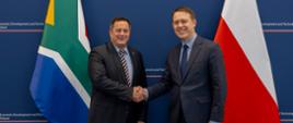 From the left, against the background of the flags of South Africa and the Republic of Poland, standing: Deputy Minister of Trade, Industry and Competition of South Africa, John Steenhuisen, and Deputy Minister of Development and Technology, Michał Baranowski; a Ministry of Trade, Industry and Competition backdrop is visible in the background; the politicians are shaking hands.