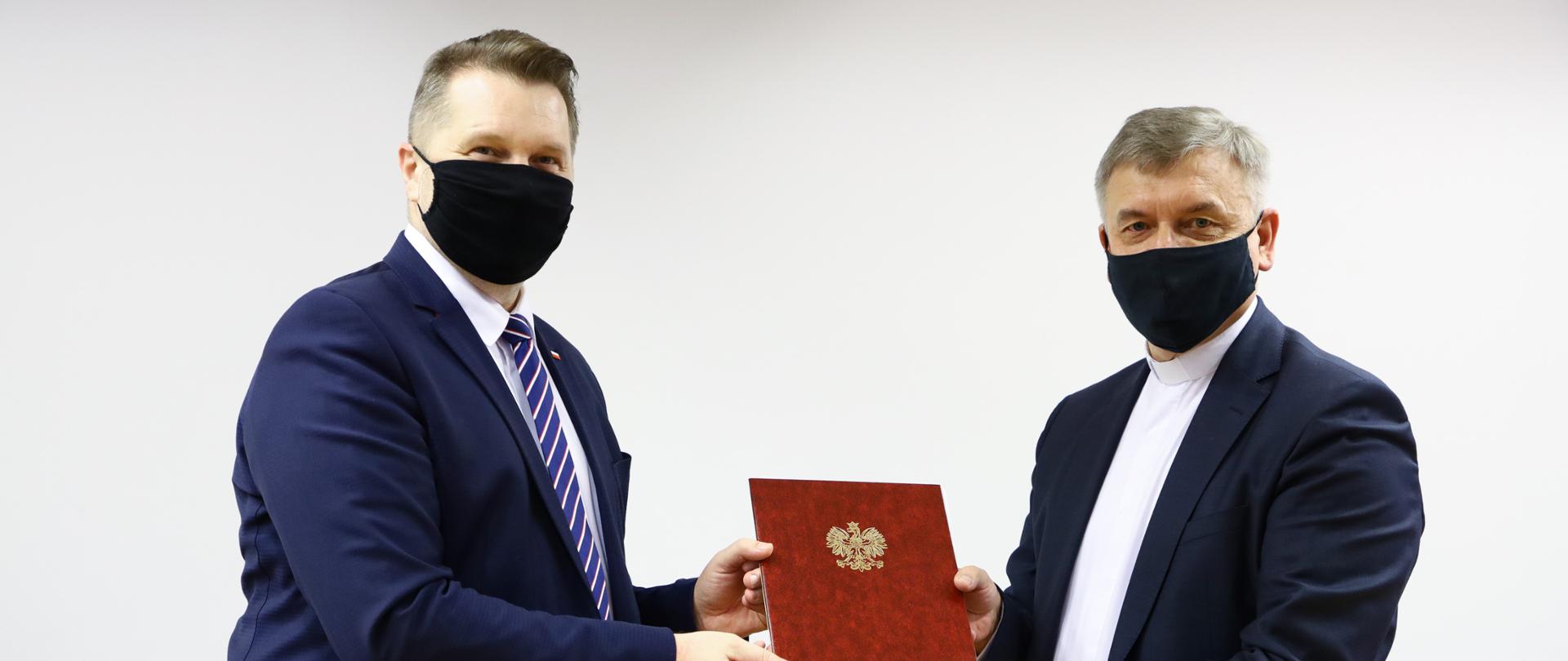 Minister Przemysław Czarnek i rektor UKSW ks. prof. dr. hab. Ryszard Czekalski stoją trzymając w rękach umowę.