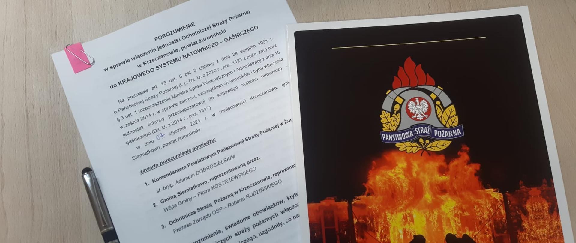 Na drewnianym stole leży teczka czarno- czerwona z logiem PSP spod niej wystaje biała kartka z czarnymi napisami. Obok kartki leży długopis.