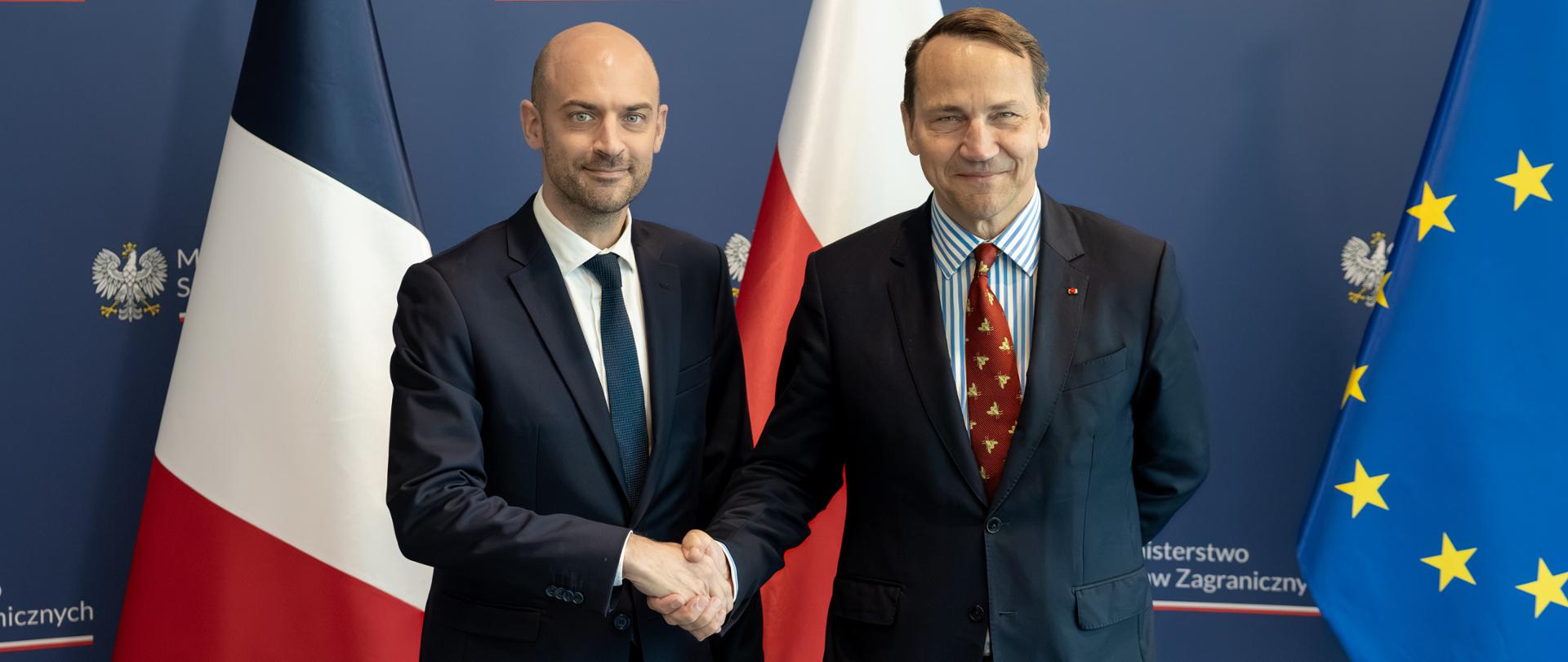 The photo shows two men in suits. They are shaking hands. They are looking toward the camera. Behind them is a wall with the words "Ministry of Foreign Affairs" and two flags—those of France and Poland. 