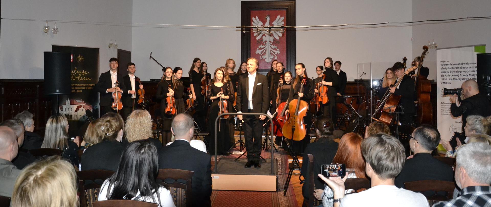 zdjęcie na pierwszym planie przedstawia publiczność zgromadzoną w sali królewskiej PSM Mielec na koncercie charytatywnym dla Klary, dalej znajduje się orkiestra smyczkowa z dyrygentem Ryszardem Kuskiem na przedzie, w tle widać fragment godła polski i portretów władców polski