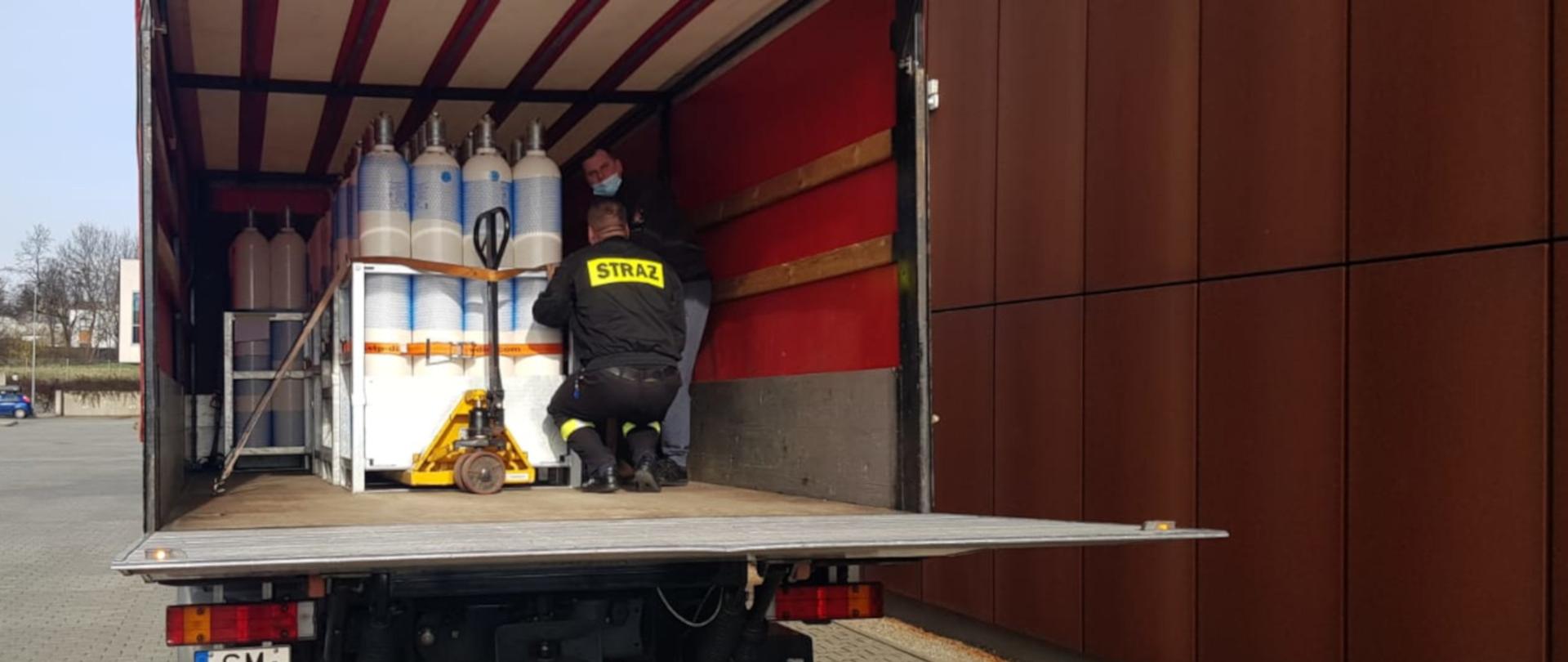 Na zdjęciu widoczny jest parking przy KM PSP w Mysłowicach. Na pierwszym planie samochód kwatermistrzowski PSP, na którym w przestrzeni ładunkowej znajdują się butle z tlenem medycznym. W samochodzie znajduje się dwóch mężczyzn w tym strażak w ubraniu koszarowym. 