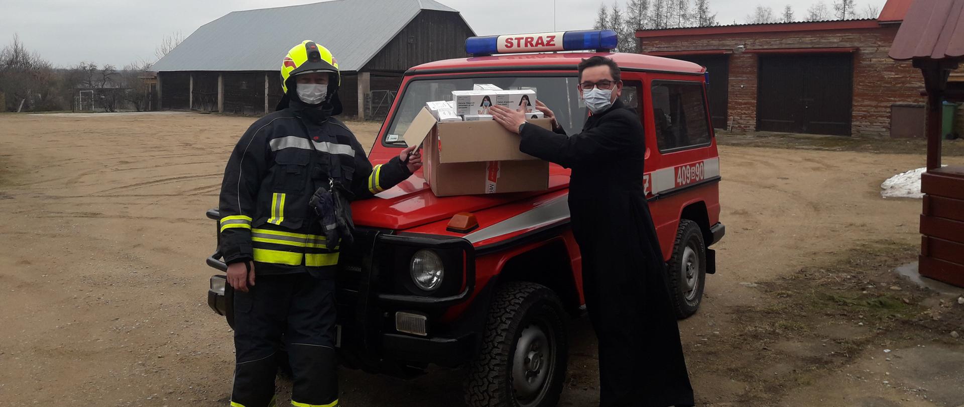 Na zdjęciu widać strażaka przekazującego maseczki ochronne dla Księdza.