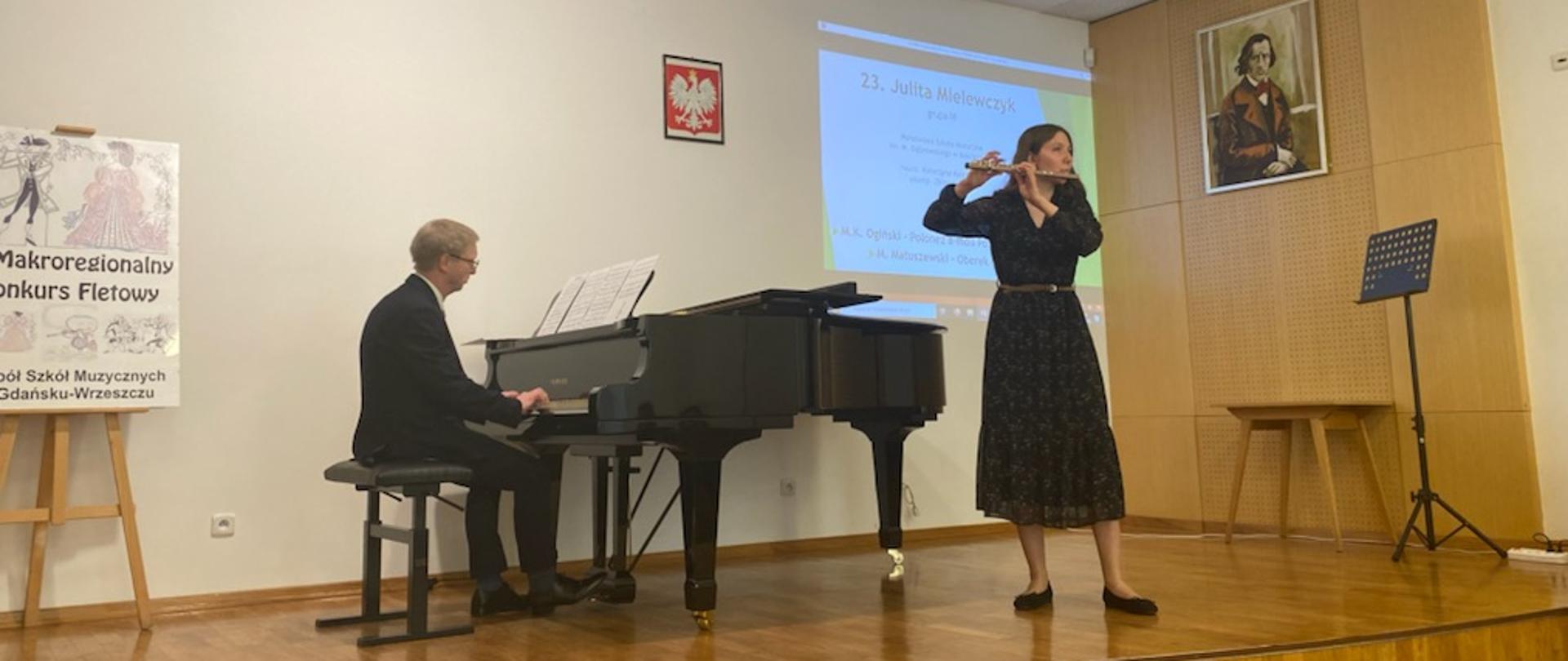 Zdjęcie przedstawia występ uczennicy podczas konkursu. Uczennica gra na flecie. na scenie stoi fortepian, a przy nim akompaniator. Scena pokryta parkietem. Ściany w kolorze kremowym. Na scenie stoi też mały stolik, pulpit oraz plakat imprezy. Na ścianach godło i obraz.