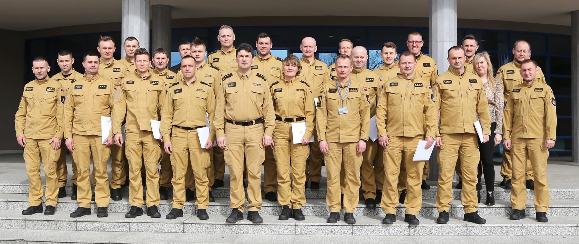 Grupowe zdjęcie uczestników szkolenia (20 osób) w towarzystwie Komendanta Centralnej Szkoły PSP, Zastępcy Komendanta Centralnej Szkoły PSP, trzech oficerów CS PSP oraz kobiety