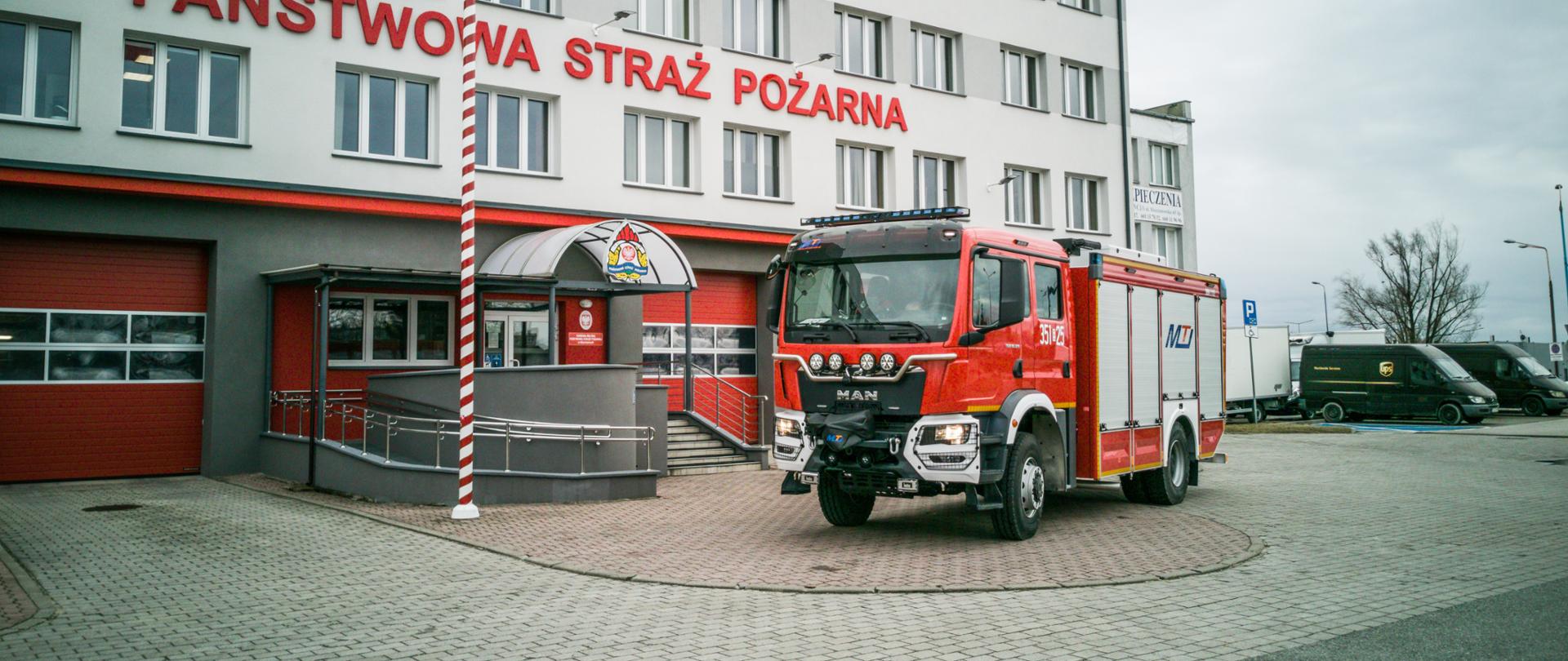 Przód i lewa strona samochodu pożarniczego w kolorze czerwonym marki MAN. W tle ściana budynku komendy i maszt z flagą polski.