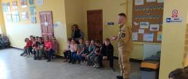 Na korytarzu szkolnym funkcjonariusz PSP prowadzi pogadankę dla dzieci siedzących na ławkach.