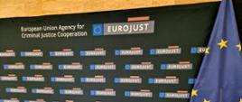 Logo EUROJUST (biały napis na szarym tle) z flagą Unii Europejskiej (okręg z żółtych gwiazd na niebieskim tle) 