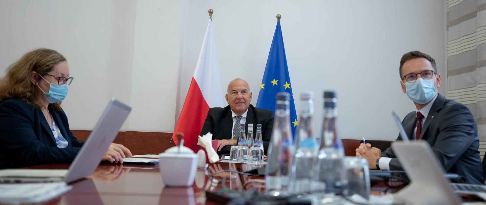 Minister Kościński, minister Buda, i dyrektor Kusina siedzą przy stole, na którym stoją komputery, za nimi stoi flaga Polska i UE 