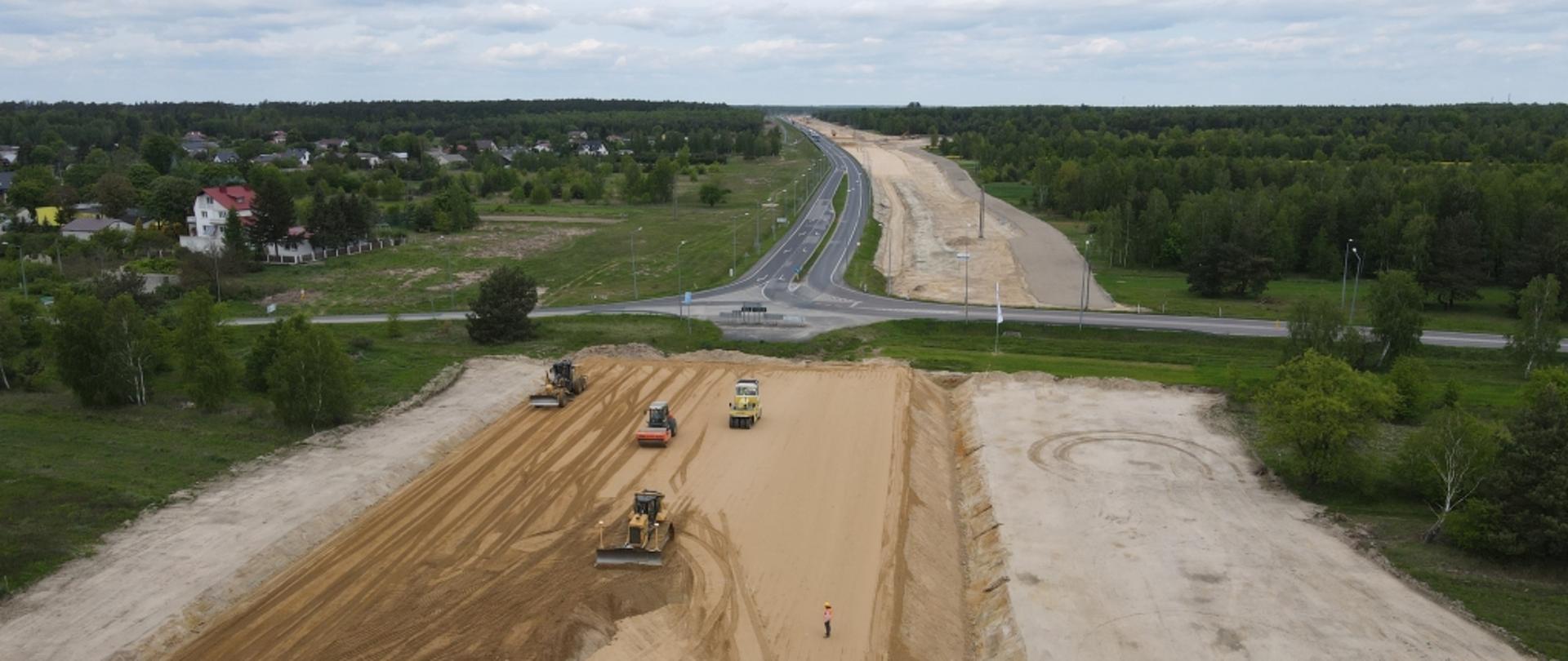Teren budowy drogi ekspresowej, widoczne maszyny budowlane, po obu stronach budowanej drogi tereny zielone, po lewej stronie dodatkowo budynki mieszkalne.