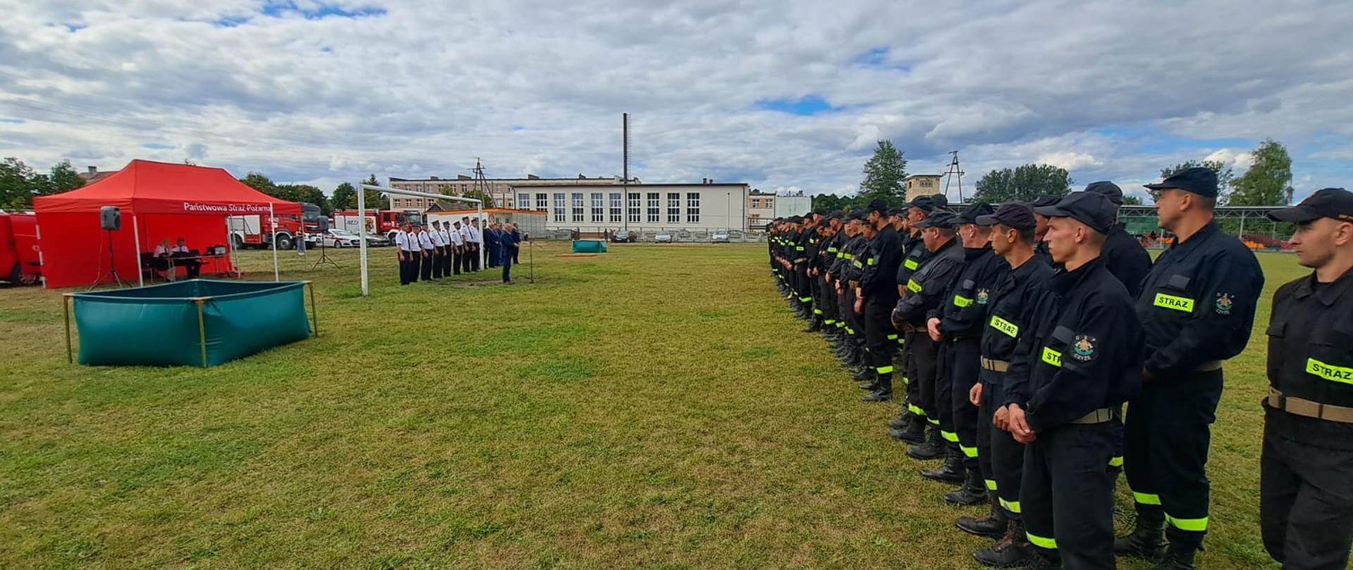 Na boisku w dwuszeregu stoi kilkudziesięciu strażaków. W oddali czerwony namiot.