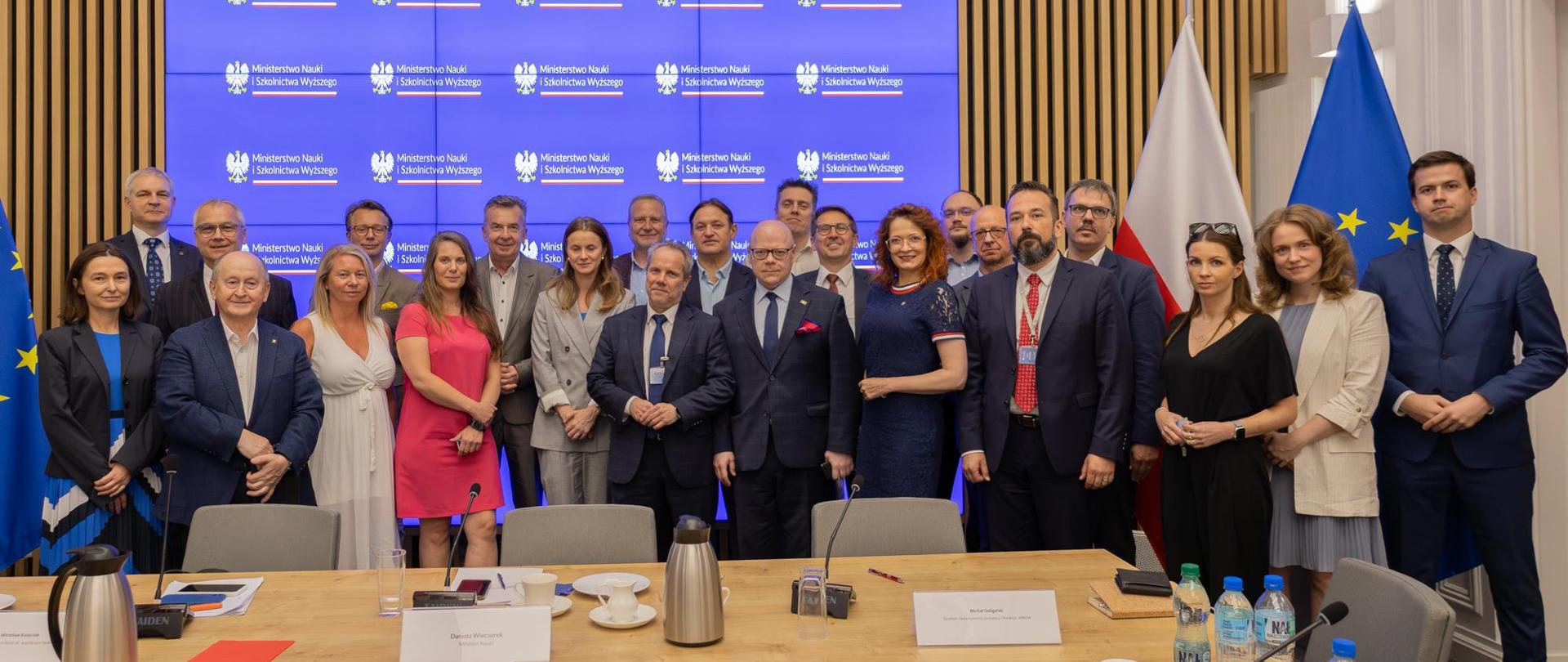 Zdjęcie zbiorowe, pod wbudowanym w ścianę sali wielkim ekranem stoi duża grupa osób, pośrodku minister Wieczorek.