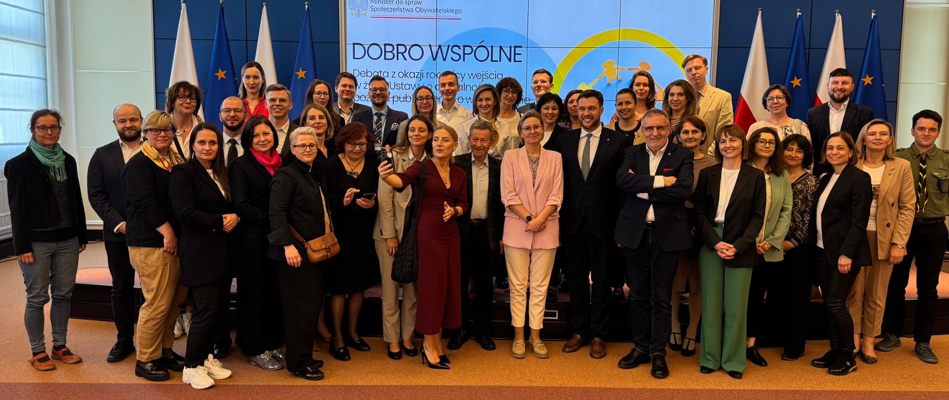 Zdjęcie grupowe pokazuje uczestników wydarzenia - Debata Dobro Wspólne - 24 kwietnia 2025 r. w Kancelarii Prezesa Rady Ministrów, stojących wspólnie na tle ekranu, na którym wyświetlany jest tytuł debaty. W tle widoczne są również flagi Polski i Unii Europejskiej.