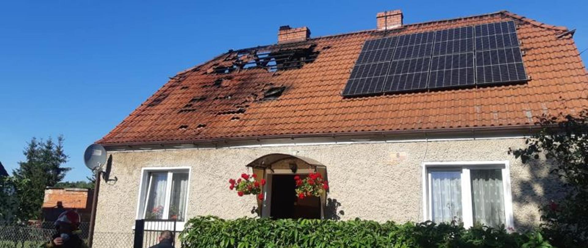 Zdjęcie wykonane po ugaszeniu pożaru. Przedstawia budynek po pożarze. Na dachu brakuje dachówek. Jest w nim dziura. Dachówki przy dziurze okopcone. Dach z pomarańczowych dachówek.