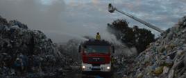 Pożar na składowisku odpadów. Plac składowy jednej z firm zajmującej się m.in. przetwarzaniem odpadów. Pomiędzy dwiema hałdami stoi wóz straży pożarnej, na jego dachu stoi strażak, który gasi pożar przy użyciu działka samochodowego. Z prawej strony, nad hałdy wysunięty jest kosz drabiny mechanicznej. W koszu jest strażak prowadzący działania gaśnicze. Panuje silne zadymienie.