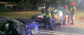 dwa samochody z rozbitymi przodami na drodze jeden koloru granatowego drugi srebrny stoją na drodze obok samochodu srebrnego stoją strażacy i policjant w oddali widać drzewa 