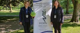 Dwie kobiety w bluzach z logo PIORiN stoją przy na wolnym powietrzu przy banerze z napisem Wojewódzki Inspektorach Ochrony Roślin i Nasiennictwa w Olsztynie