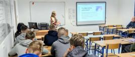 Uczniowie siedzący w ławkach słuchają prelekcji prowadzonej przez kobietę. Na tablicy wyświetlona jest prezentacja.