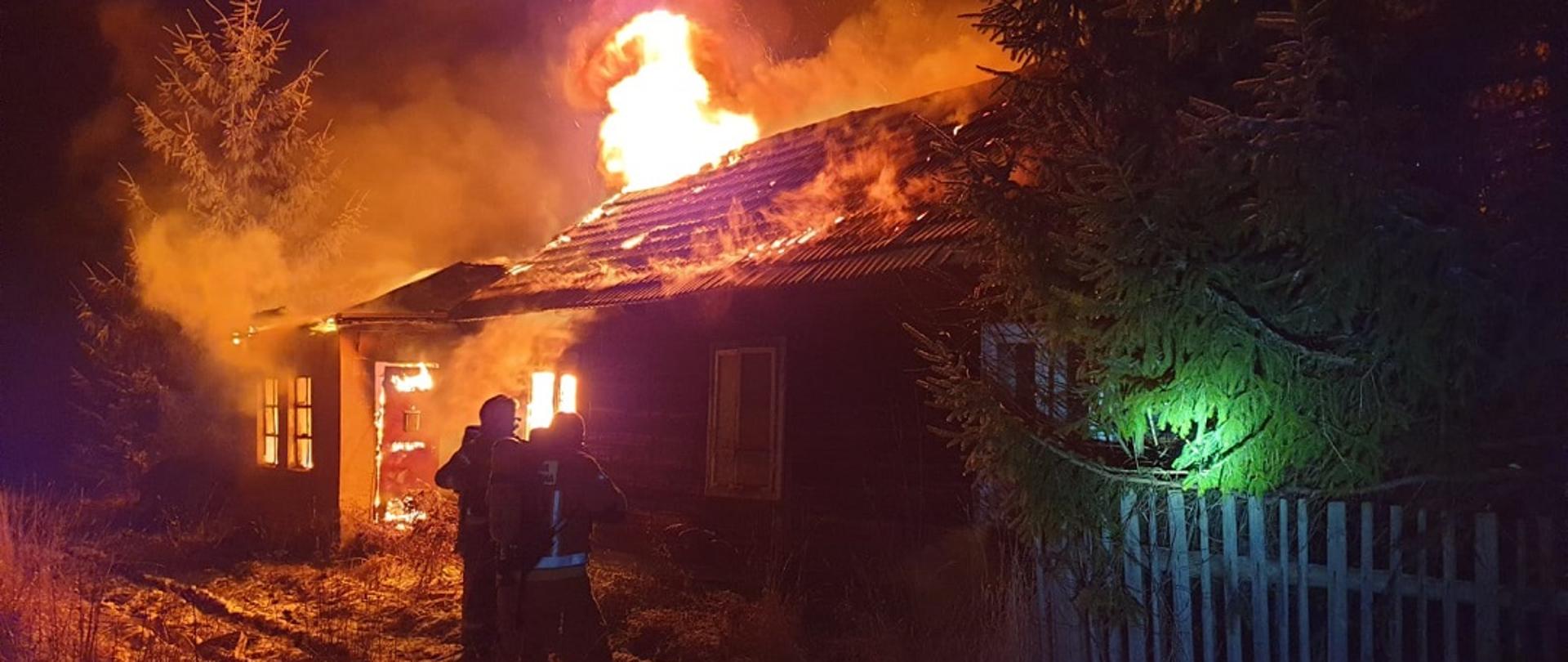 Zdjęcie obrazuje budynek murowano-drewniany w kształcie litery C. Pożar całkowicie objął budynek. Płomienie widać wewnątrz obiektu, jak i na zewnątrz. Z dachu wydobywa się pełno dymu oraz miejscami jęzory ognia. Przed obiektem dwóch strażaków ubranych w obrania bojowe, hełmy i zabezpieczonych aparatami ochrony dróg oddechowych podaje prąd wody na palący się budynek. 