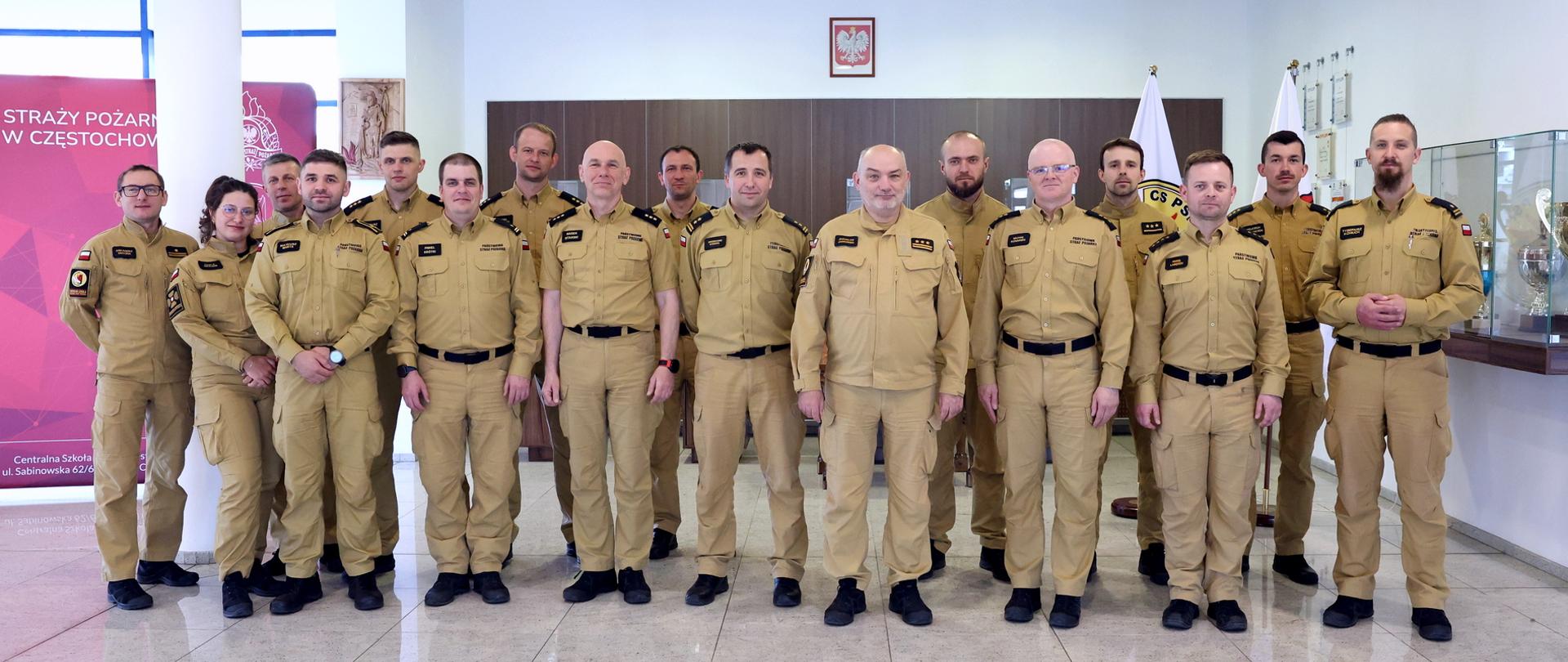 Grupa strażaków Państwowej Straży Pożarnej w piaskowych mundurach służbowych stoi do wspólnego zdjęcia w pomieszczeniu Centralnej Szkoły PSP w Częstochowie.
