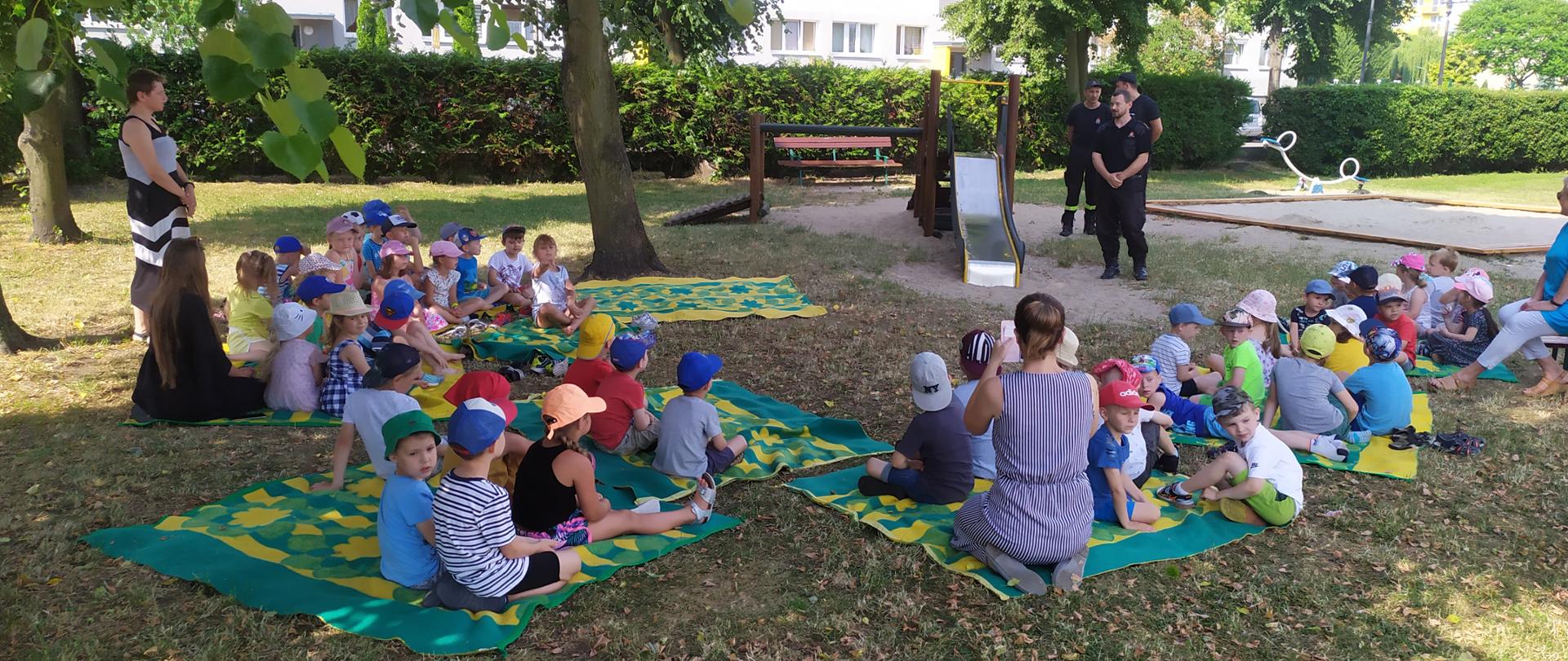 Zdjęcie przedstawia dzieci siedzące na rozłożonych na trawie kocach. Przedszkolaki z Przedszkola im. Kubusia Puchatka w Ciechocinku wraz z wychowawczyniami podczas pogadanki prowadzonej przez strażaków z JRG Ciechocinek. Na drugim planie trzech strażaków, plac zabaw i drzewa.