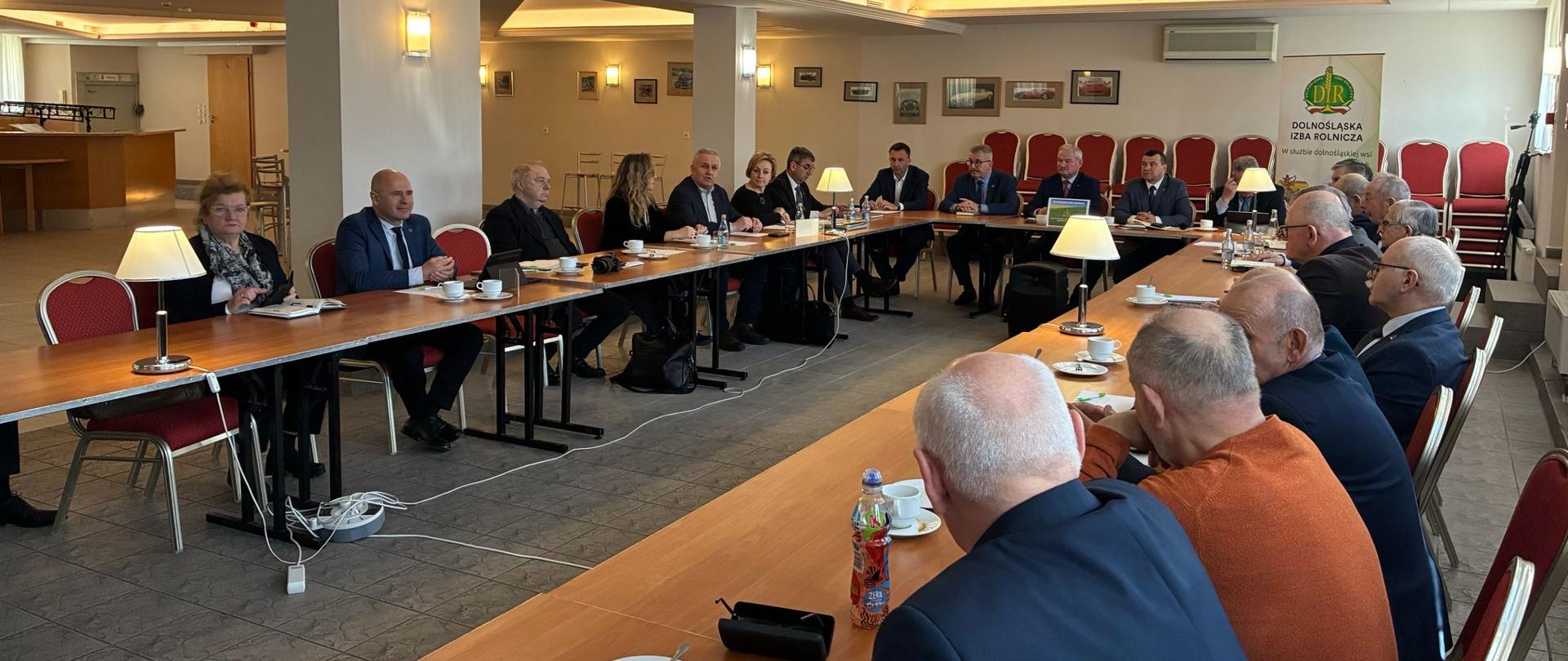 Uczestnicy spotkania siedzą przy stołach ustawionych w podkowę w sali konferencyjnej