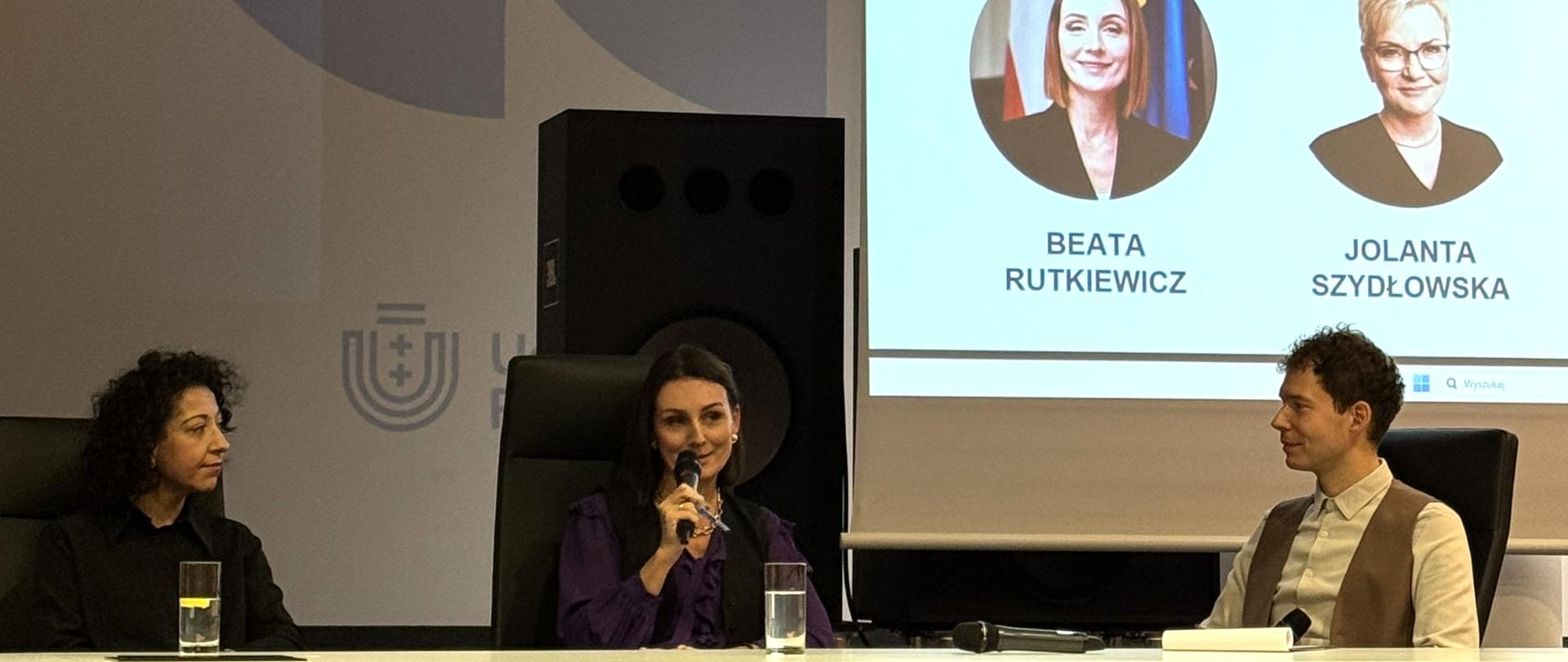 kobieta siedzi za stołem i przemawia przez mikrofon 