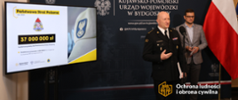 Strażak w mundurze wyjściowym stoi przy mikrofonie, za nim mężczyzna w marynarce. Z lewej strony widać ekran z informacją, a w tle ścianka reklamowa Urzedu. Z prawej strony flaga biało-czerwona.