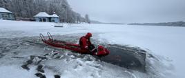 Strażacy w czerwonych ubraniach wypornościowych do pracy w wodzie ćwiczą podejmowanie osoby poszkodowanej, pod którą załamał się lód i wpadła do wody. Na zdjęciu strażacy na saniach lodowych. Zamarznięte jezioro w zimowej aurze. Strażak podejmuje poszkodowanego z wody.